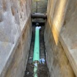 New green sewer line being installed at the bottom of a narrow trench